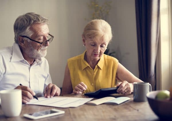 Ouder echtpaar waarbij de vrouw misprijzend kijkt naar de rekenmachine, maar de man kijkt net hoopvol op van zijn papieren.