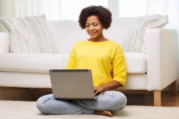 Vrouw in gele trui zit tegen een witte zitbank aan met laptop op schoot